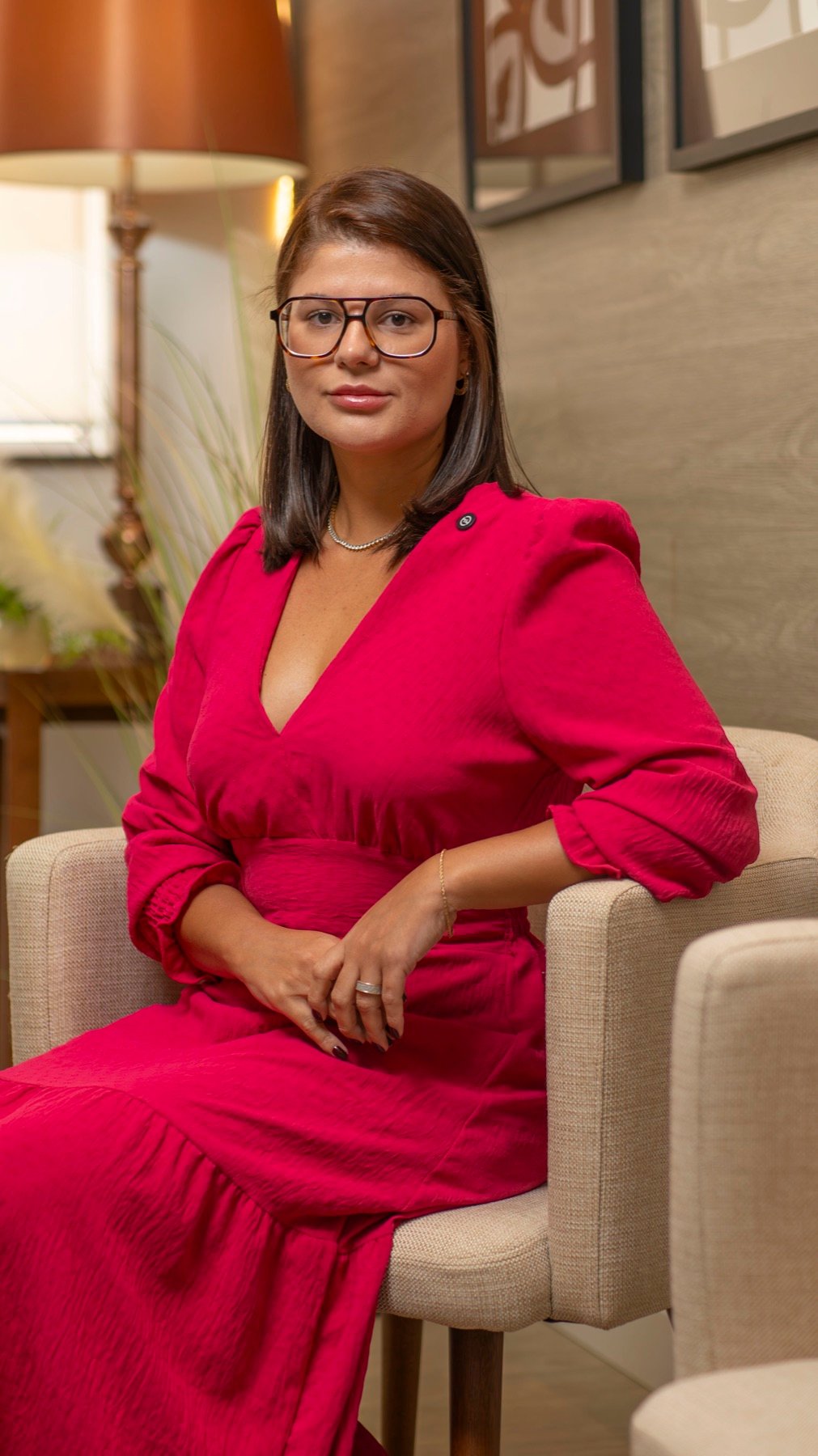 Dra. Ludmila Barbosa sentada em poltrona da clínica, vestindo vestido vinho, em pose acolhedora e elegante